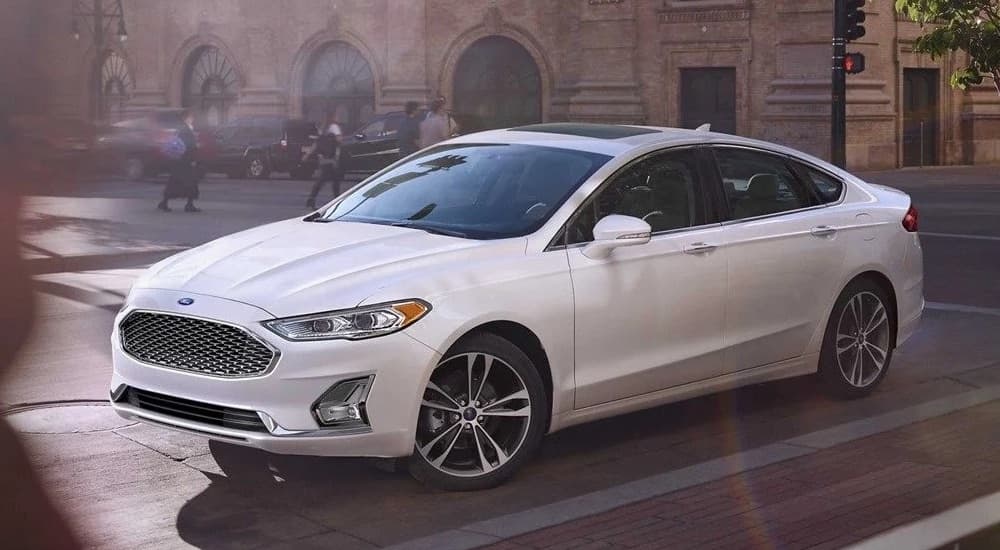 A white 2017 Ford Fusion driving on a city street near a used car dealership.
