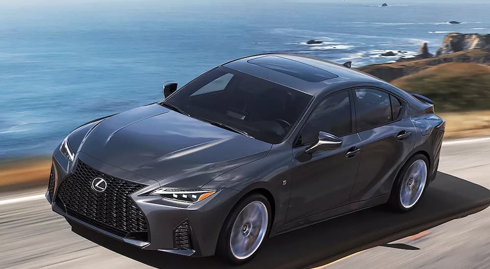 A grey 2022 Lexus IS is shown driving on a highway next to an ocean.